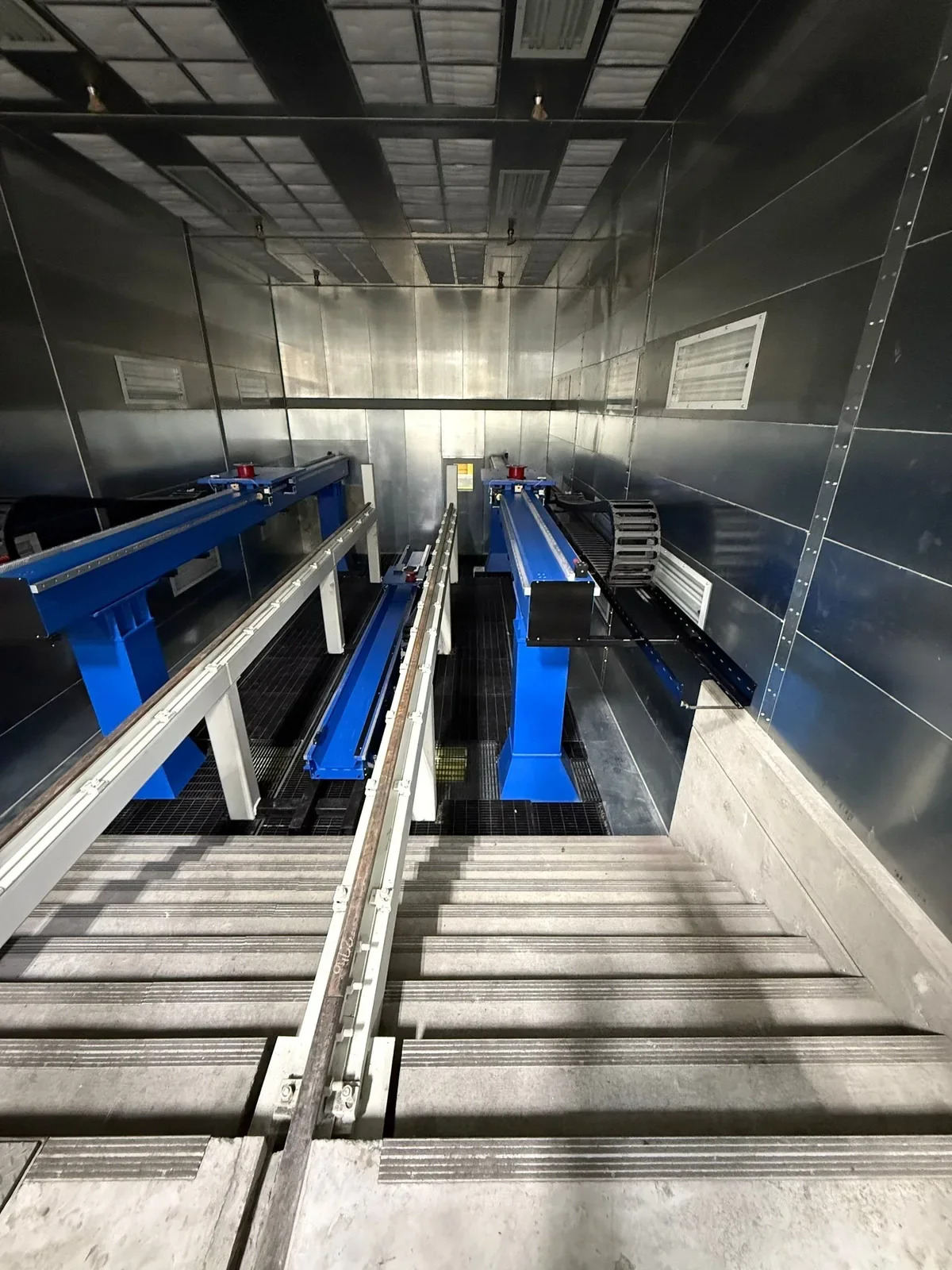 Blue handling equipment and parallel rails within a metal booth, viewed down a sloped floor opening.