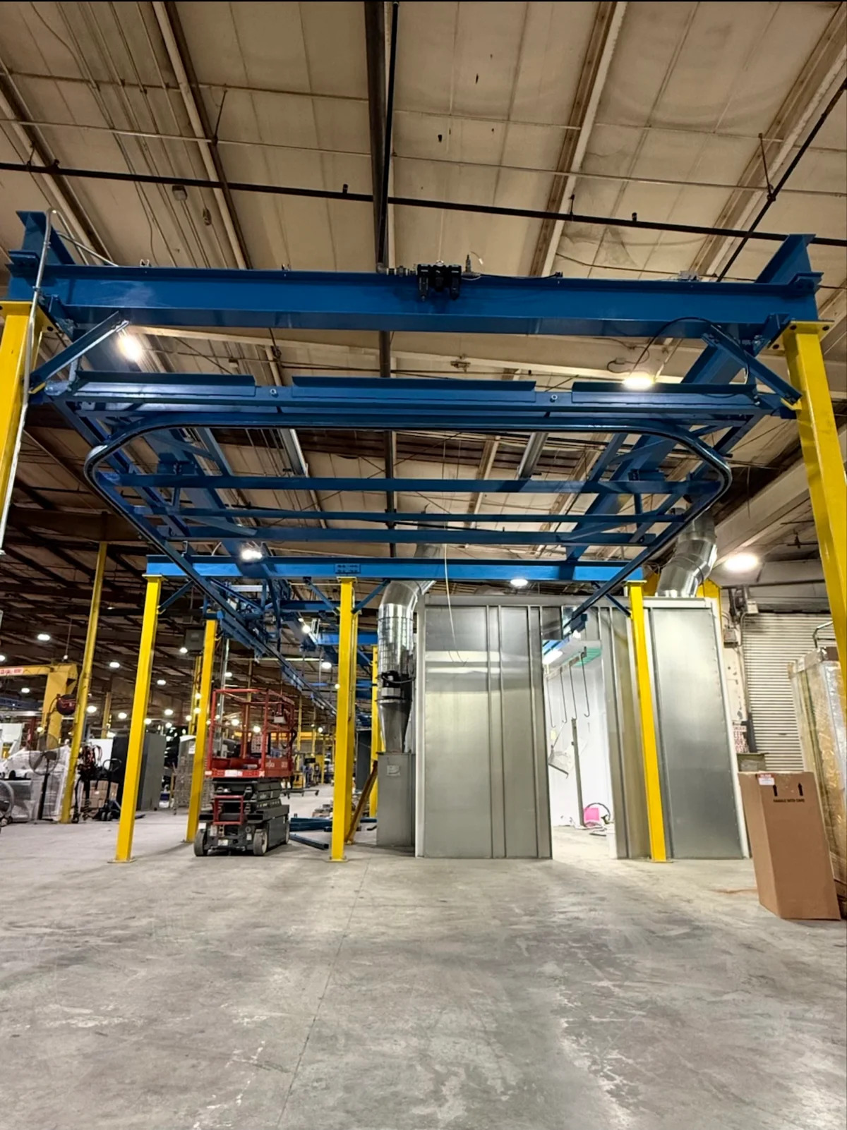 Front view of blue curved overhead conveyor track and supporting beams on yellow columns beside finishing equipment.
