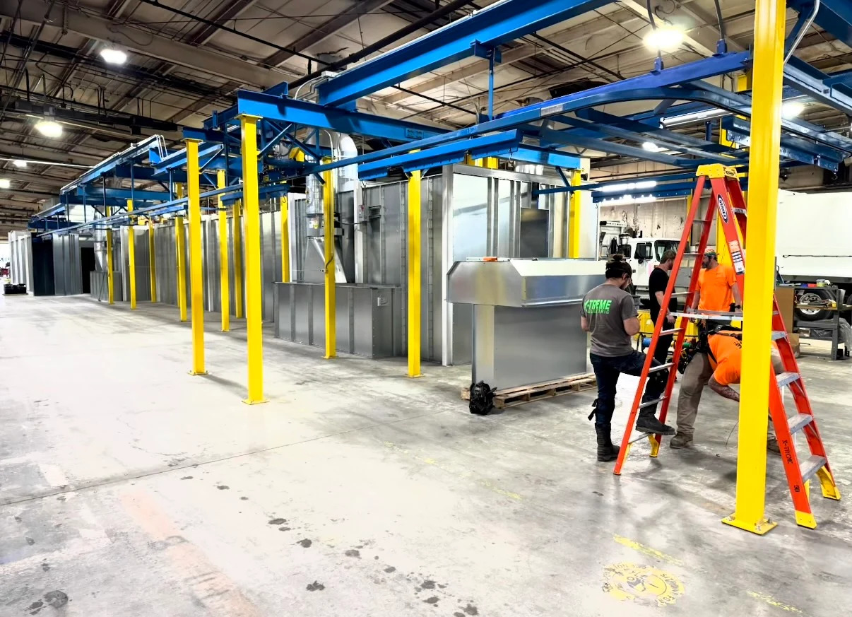 Wide view of blue overhead conveyor routing and yellow support columns beside finishing equipment during installation.
