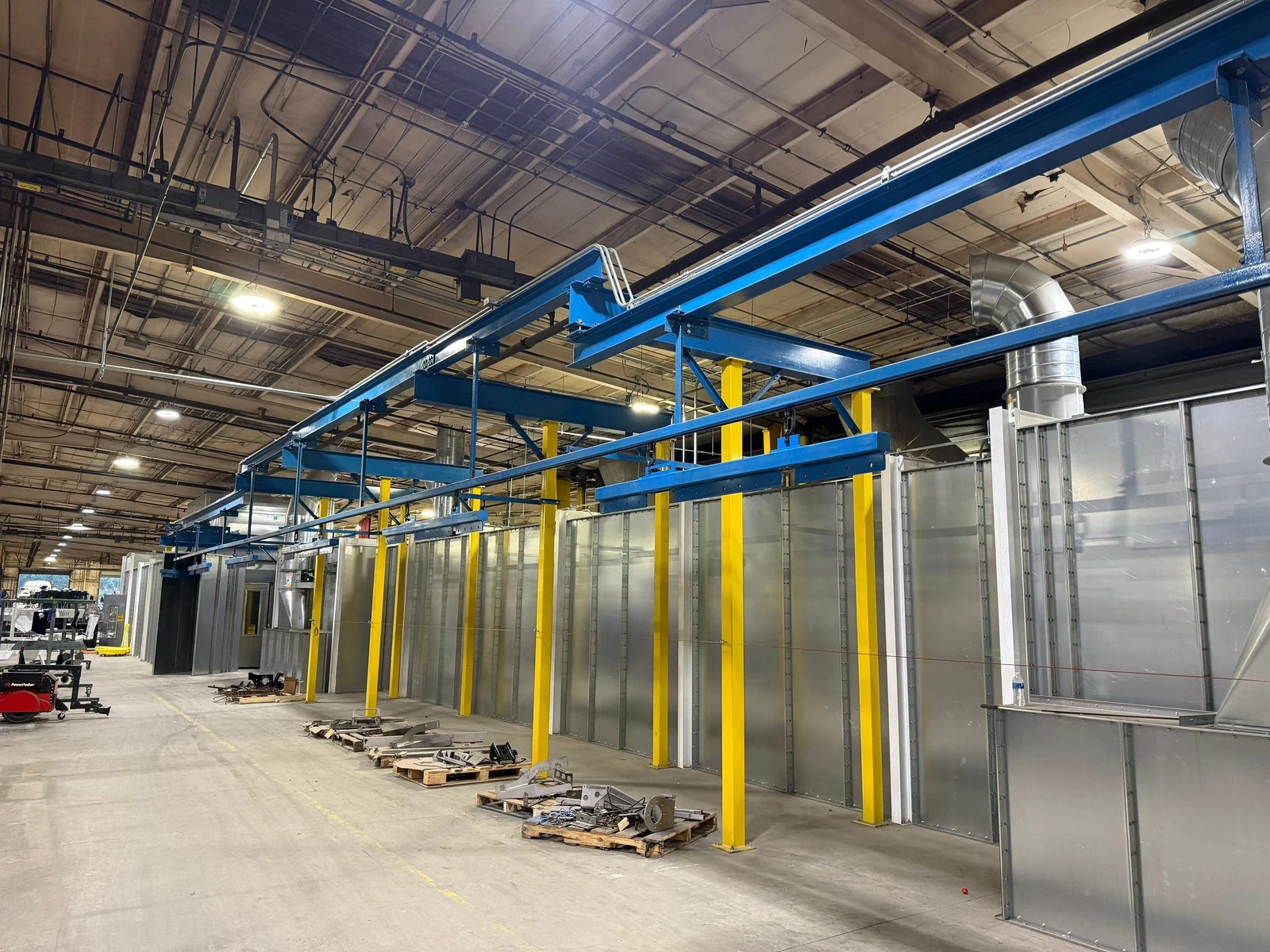Blue overhead conveyor track and support beams on yellow columns beside finishing equipment and staged installation materials.