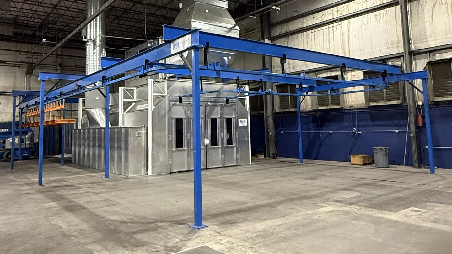 Hand-pushed conveyor route around a finishing booth, with blue freestanding support steel and orange load bars