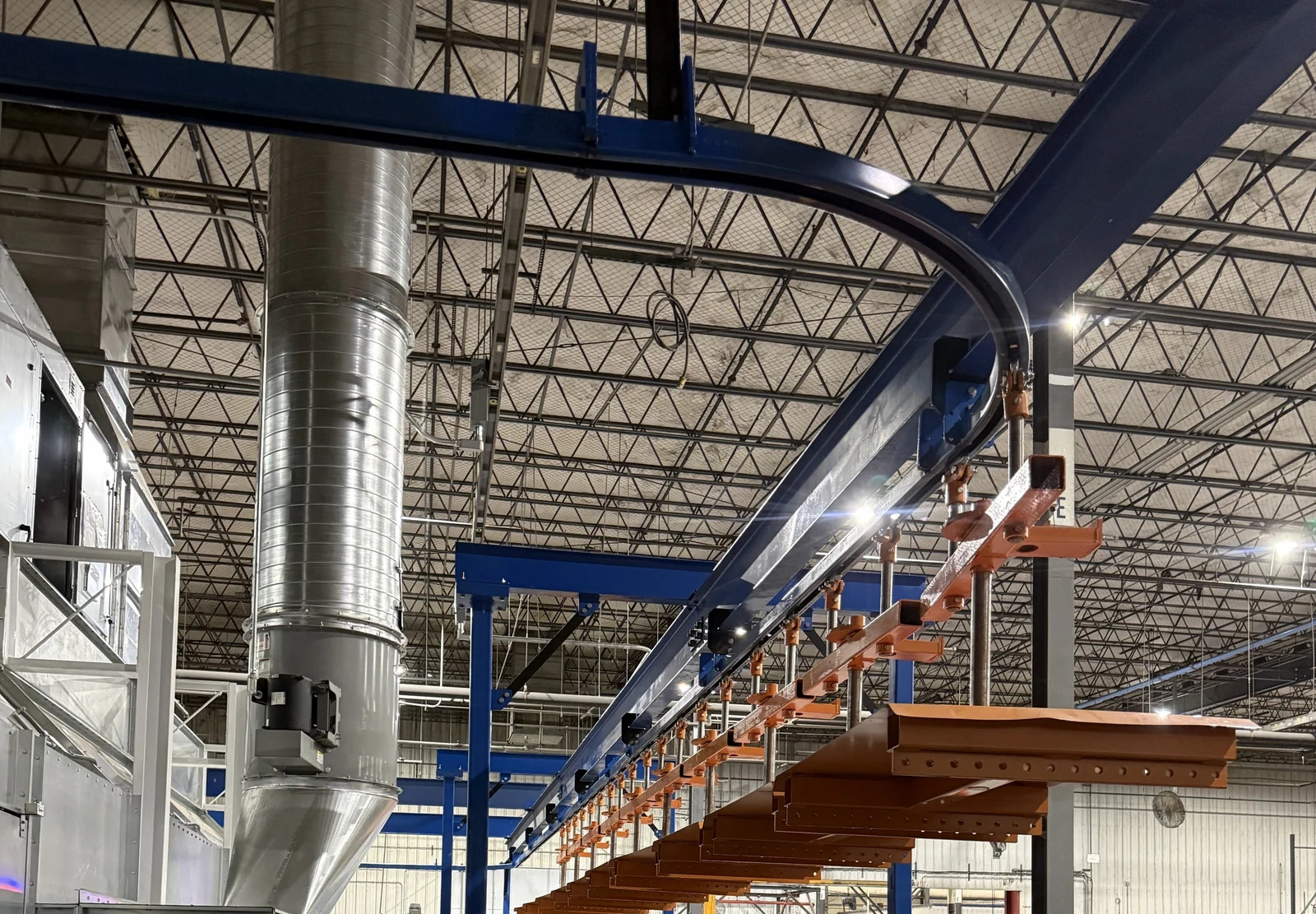 Orange hand-pushed trolleys and load bars following blue overhead track beside a finishing booth.